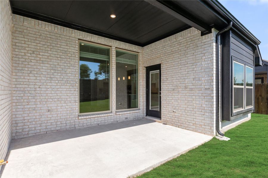Covered back patio with concrete slab, light brick exterior, large windows, and black door leading inside