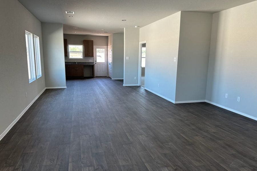 Image of home living areas with kitchen in rear. Wood-look flooring, white walls and windows down one wall, on rear door, and over sink.
