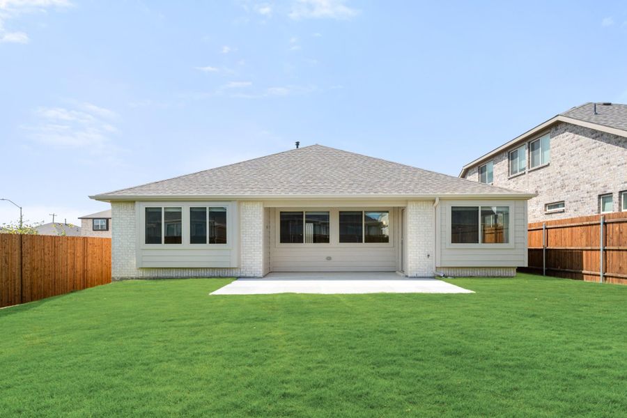 Rear view of single-story home with white brick exterior, large windows, concrete patio, and fenced green backyard Rear view of single-story home with white brick exterior, large windows, concrete patio, and fenced green backyard