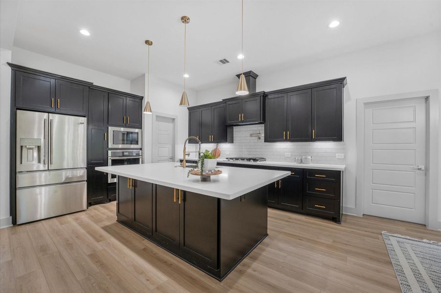 This modern kitchen features sleek black cabinetry with gold handles, a large island with a white countertop, and stainless steel appliances. It has contemporary pendant lighting and a spacious layout with light wood flooring. This modern kitchen features sleek black cabinetry with gold handles, a large island with a white countertop, and stainless steel appliances. It has contemporary pendant lighting and a spacious layout with light wood flooring.