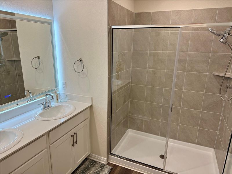 Spacious bathroom featuring a dual vanity with white cabinetry, a light-toned countertop, chrome fixtures and walk-in shower.