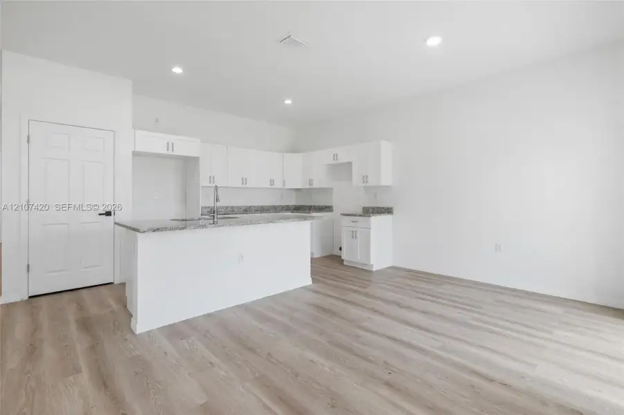 Kitchen featuring white shaker-style cabinetry, stone-finish countertops, an island with an integrated stainless steel faucet, recessed lighting, and wood-finish flooring Kitchen featuring white shaker-style cabinetry, stone-finish countertops, an island with an integrated stainless steel faucet, recessed lighting, and wood-finish flooring