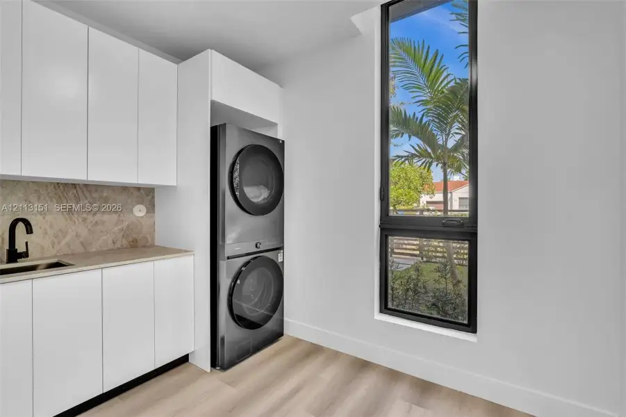 Laundry area featuring floor-to-ceiling white cabinetry, stone-finish backsplash, built-in sink with matte black faucet, stacked laundry appliances, and tall window with black frame Laundry area featuring floor-to-ceiling white cabinetry, stone-finish backsplash, built-in sink with matte black faucet, stacked laundry appliances, and tall window with black frame
