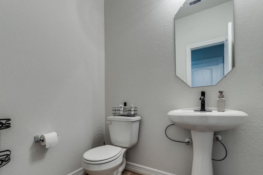 This is a compact, modern bathroom with a pedestal sink, toilet, and a geometric mirror. The walls are painted a light neutral color, and there's a simple fixture for soap and toiletries.
