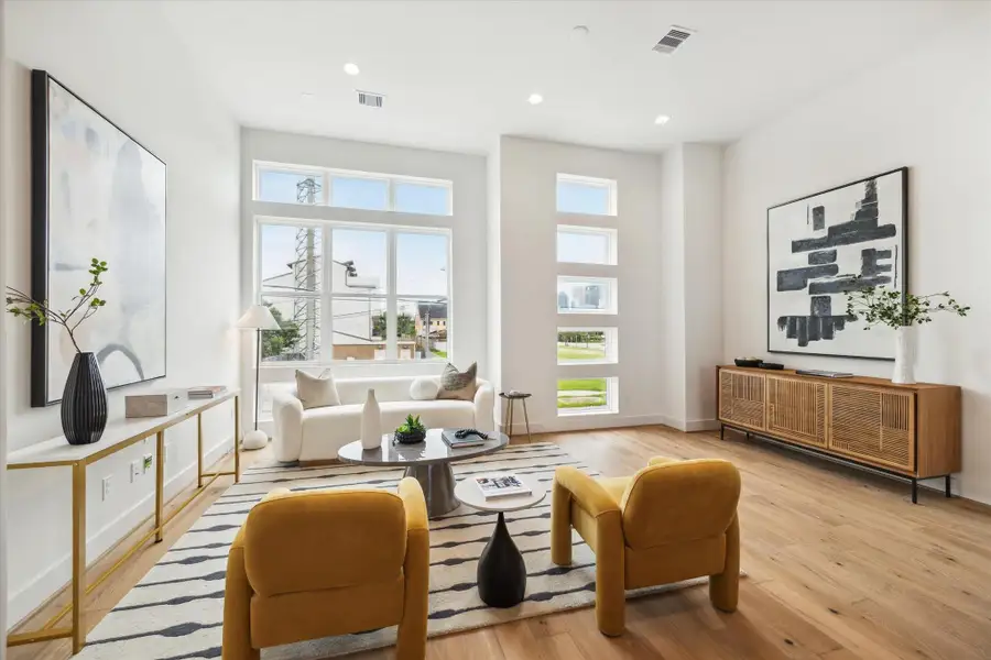 This modern living room features large windows that provide ample natural light, light wood flooring, and minimalist decor. It includes a cozy seating area with a white sofa, mustard yellow chairs, and stylish art pieces on the walls. The room is bright and inviting, perfect for relaxation and entertaining.