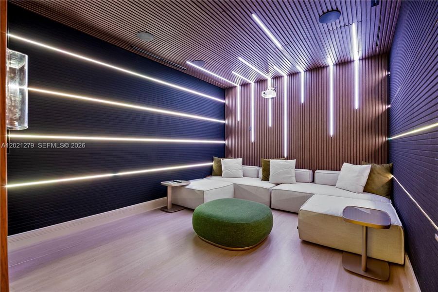 Private home theater with walnut slatted acoustic wall and ceiling panels, programmable LED linear lighting, ceiling-mounted projector, modular white sectional seating, round upholstered ottoman, and light wood floors.