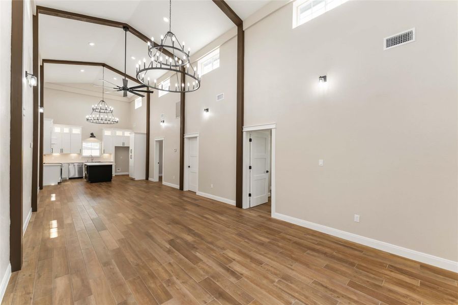 This spacious living area features high vaulted ceilings with elegant chandeliers and abundant natural light. The open layout showcases beautiful tile wood-style floors, leading into a modern kitchen with white cabinetry and a central island. Perfect for entertaining and comfortable living.