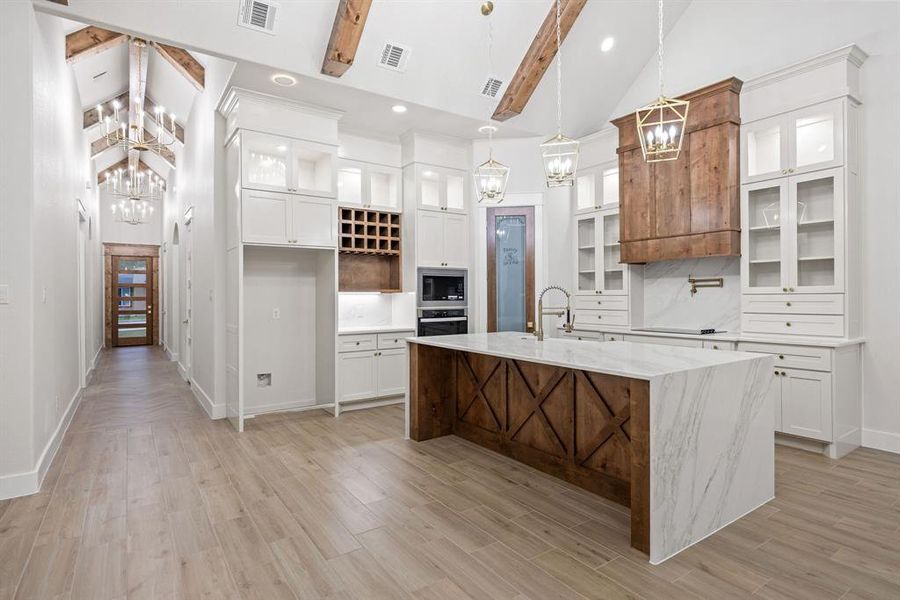 Open-concept kitchen featuring a waterfall island with a wood-paneled base and stone countertop, custom wood range hood, built-in double ovens, and a wine rack Open-concept kitchen featuring a waterfall island with a wood-paneled base and stone countertop, custom wood range hood, built-in double ovens, and a wine rack