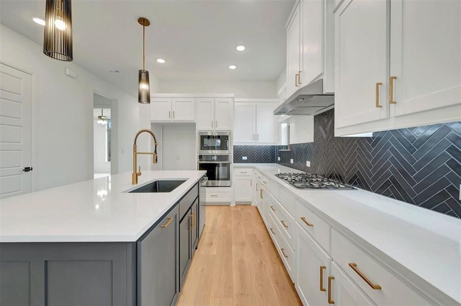 Kitchen featuring a large island with an undermount sink and a brass-finish faucet, white cabinetry, integrated stainless steel appliances, a five-burner gas cooktop, and a dark herringbone tile backsplash