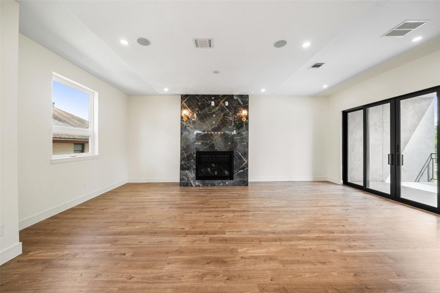 Living room features elegant wood floors, a striking dark marble fireplace, and ample natural light from large windows and glass doors, creating a bright and inviting atmosphere.