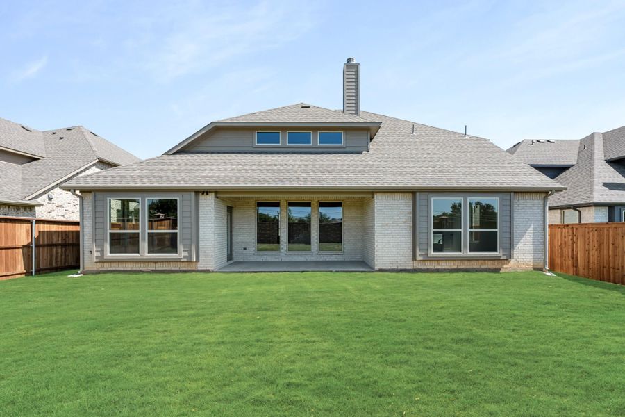 Rear exterior of single-story brick home with large windows, covered patio, chimney, and fenced backyard with green lawn