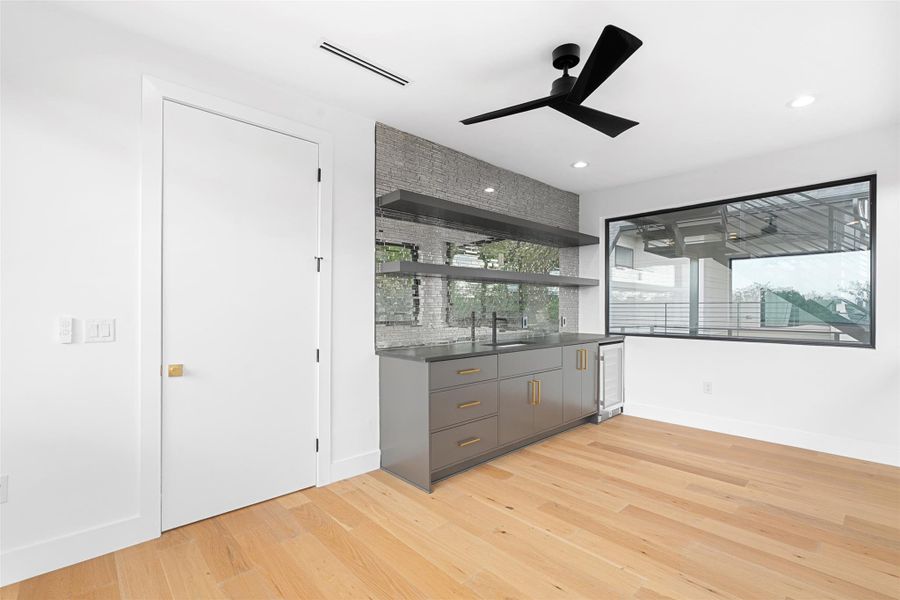 This room features light hardwood flooring, a modern ceiling fan, and a built-in wet bar with a dark countertop, sink, and an under-counter beverage cooler