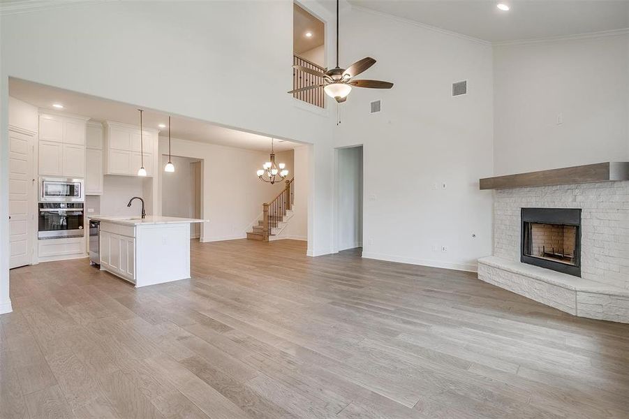 Unfurnished living room featuring ceiling fan with notable chandelier, a towering ceiling, a brick fireplace, and light wood-type flooring