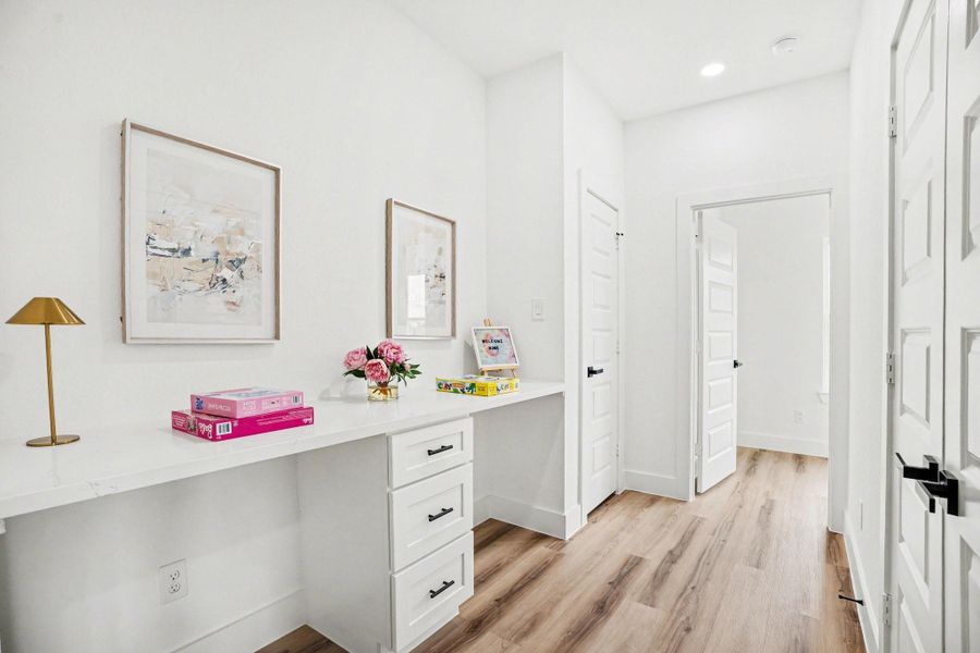 This photo showcases a bright hallway with white walls and wooden flooring. It features a built-in desk with drawers. The area is well-lit with recessed lighting.