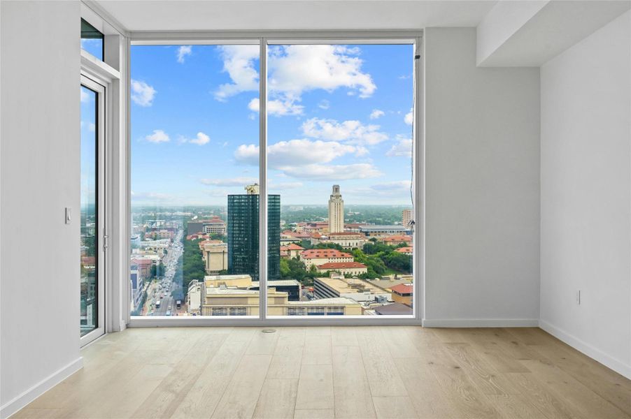 Living room with large, floor-to-ceiling glass doors leading to a balcony. The view overlooks a cityscape with prominent buildings, including the University of Texas. The room has light-colored wood flooring and a clean, modern aesthetic with a minimalist design.