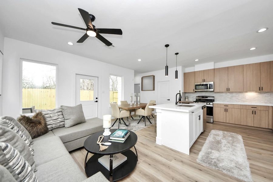 This photo showcases a modern, open-concept living area with a sleek kitchen featuring light wood cabinets, a center island, and stainless steel appliances. The space is bright and inviting, with a comfortable seating area, a dining table, and contemporary lighting. Large windows and a ceiling fan add to the airy feel. This photo showcases a modern, open-concept living area with a sleek kitchen featuring light wood cabinets, a center island, and stainless steel appliances. The space is bright and inviting, with a comfortable seating area, a dining table, and contemporary lighting. Large windows and a ceiling fan add to the airy feel.