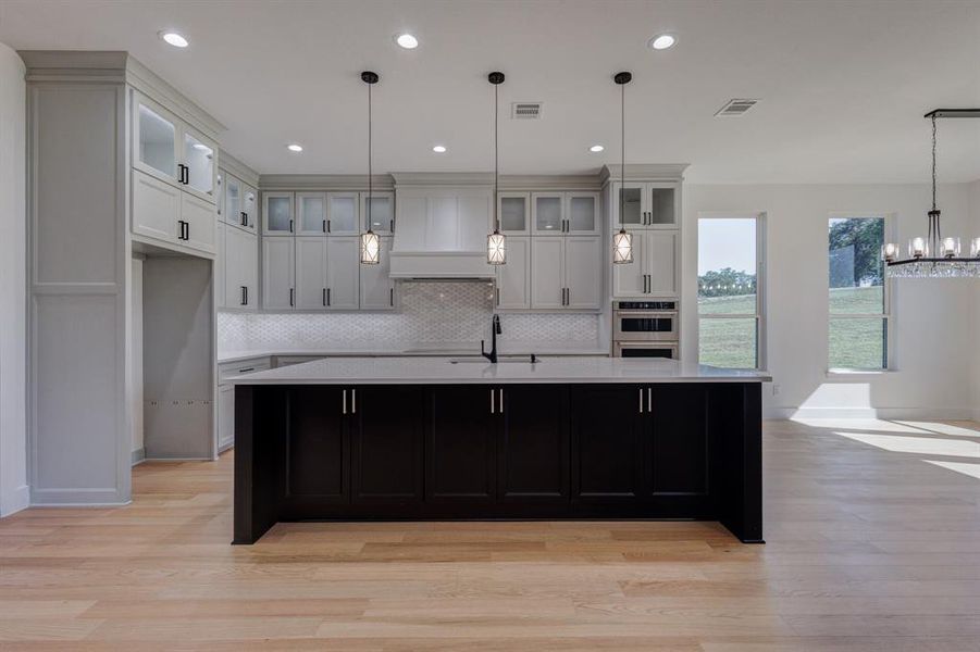 Modern kitchen featuring a large central island with dark cabinetry, light wood-finish flooring, and recessed ceiling lighting Modern kitchen featuring a large central island with dark cabinetry, light wood-finish flooring, and recessed ceiling lighting