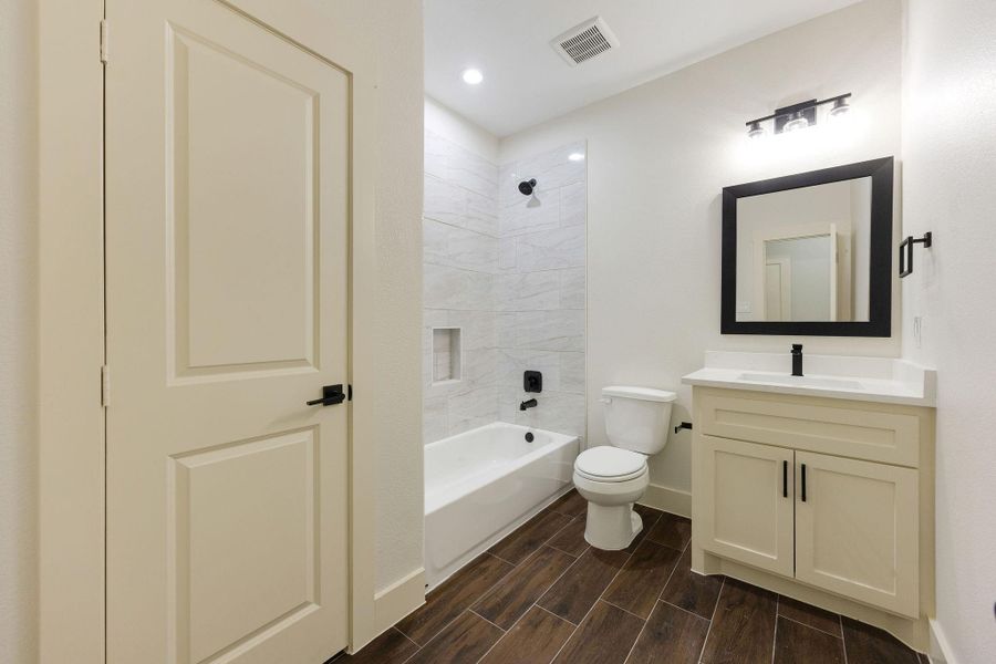 This secondary bathroom features a modern design with wood-look tile flooring, a white vanity with a black framed mirror, and a clean, white bathtub with a tiled shower. The lighting and neutral tones create a bright and inviting space.