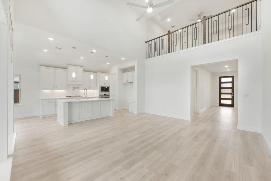 Open living area with high ceilings, light wood floors, and white kitchen with island and second-floor balcony railing visible