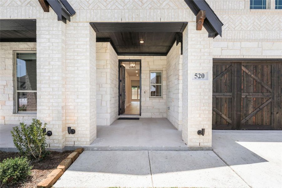 Covered front entry with light stone exterior, dark wood garage door, and open front door showing interior