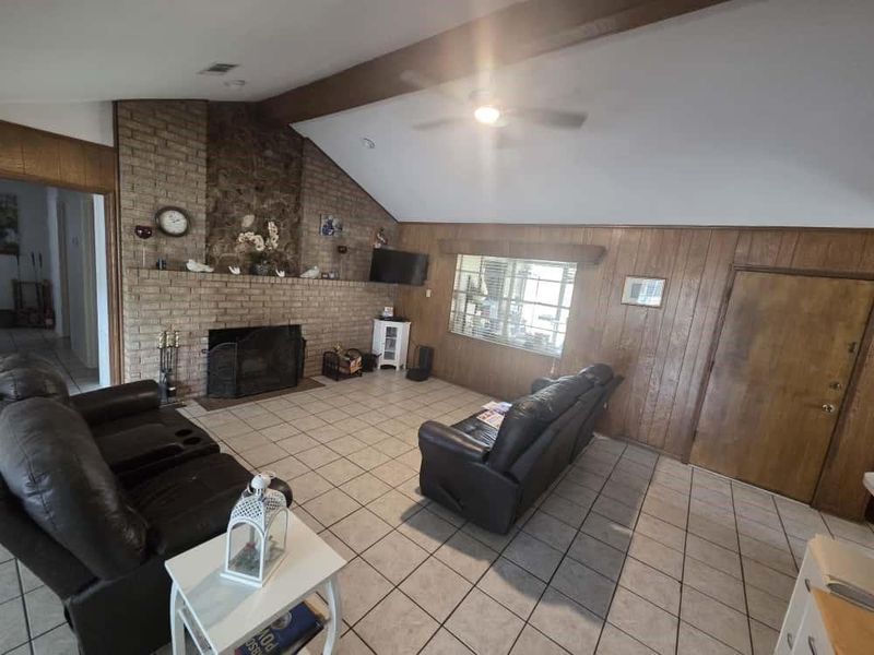 Living area featuring wood walls, light tile patterned flooring, a brick fireplace, and a ceiling fan Living area featuring wood walls, light tile patterned flooring, a brick fireplace, and a ceiling fan