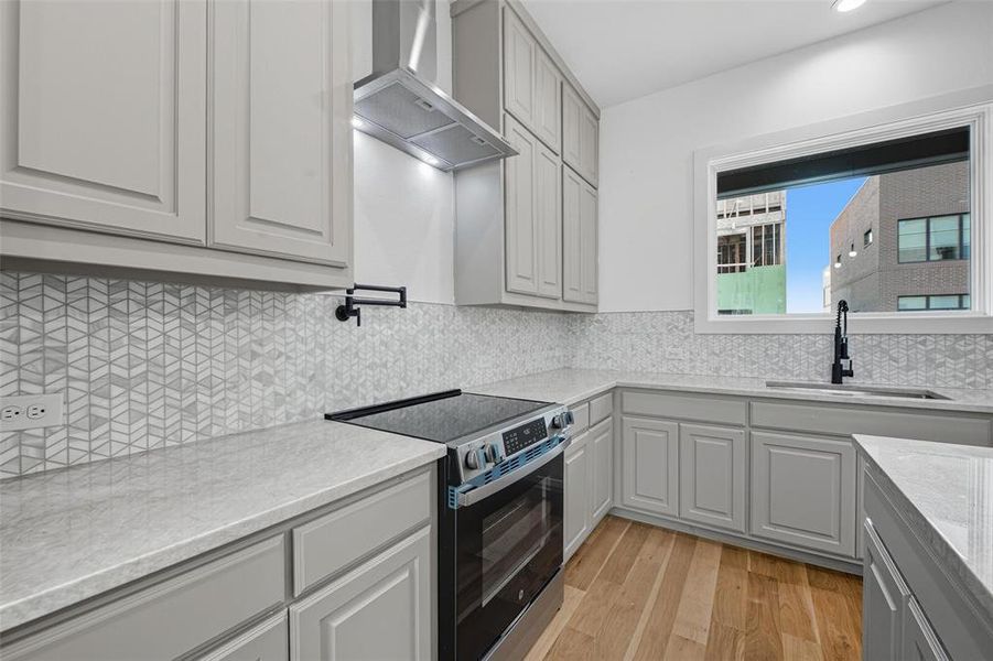 Kitchen with electric stove, gray cabinets, wall chimney exhaust hood, light wood-style floors, and decorative backsplash Kitchen with electric stove, gray cabinets, wall chimney exhaust hood, light wood-style floors, and decorative backsplash
