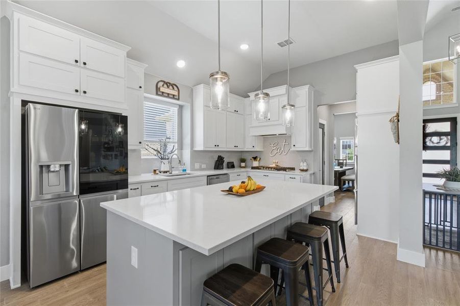 Bright kitchen featuring a central island with a light-toned countertop, white cabinetry with black hardware, stainless steel appliances, pendant lighting, and wood-finish flooring