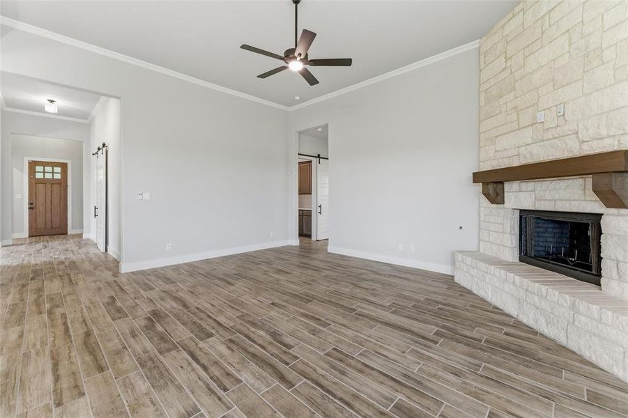 Spacious interior room featuring wood-finish flooring, a stone fireplace with a substantial wood mantel, a ceiling fan, and crown molding