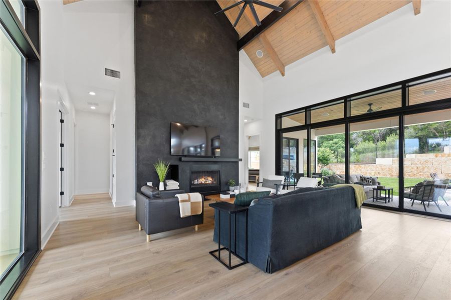 Expansive living area featuring a vaulted wood-paneled ceiling with exposed beams, a floor-to-ceiling fireplace with a dark textured finish, and wood-finish flooring Expansive living area featuring a vaulted wood-paneled ceiling with exposed beams, a floor-to-ceiling fireplace with a dark textured finish, and wood-finish flooring
