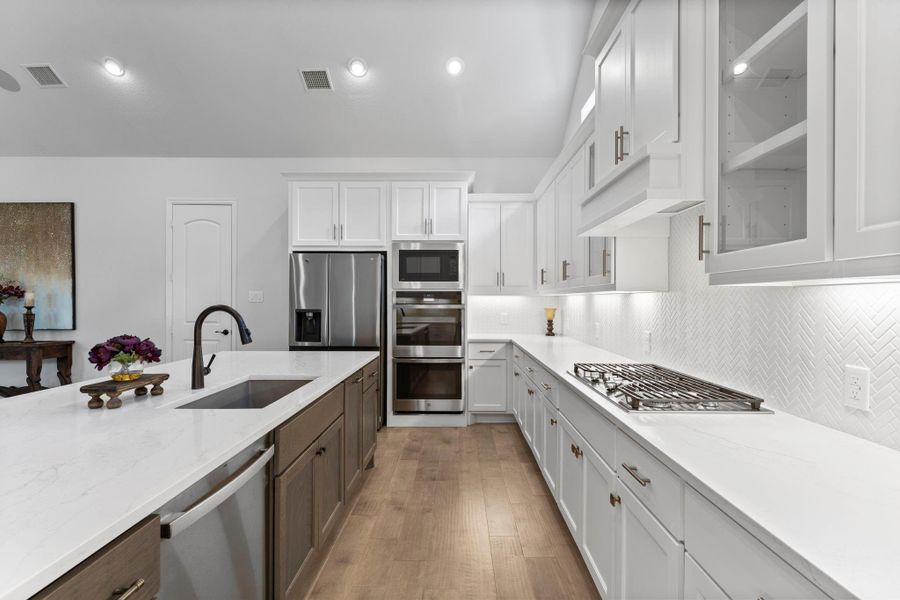 A close-up of the kitchen counter area, showing the gas cooktop and the white tile backsplash. On the left, the dark wood island with the sink and faucet is visible. The white cabinets and stainless steel appliances give the kitchen a clean, modern look. A close-up of the kitchen counter area, showing the gas cooktop and the white tile backsplash. On the left, the dark wood island with the sink and faucet is visible. The white cabinets and stainless steel appliances give the kitchen a clean, modern look.