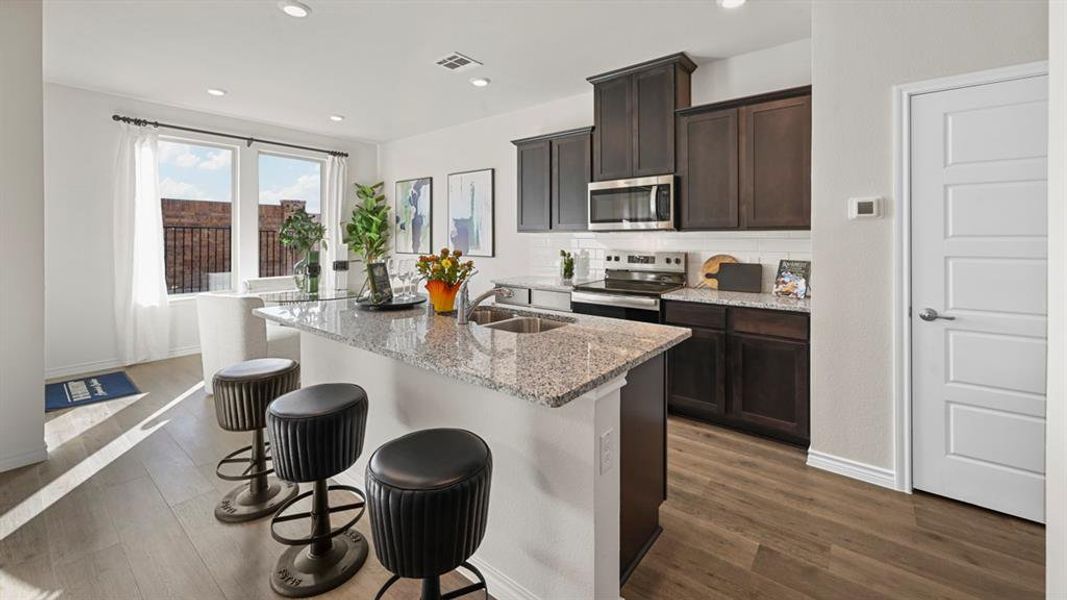 Kitchen with light stone counters, tasteful backsplash, dark brown cabinetry, stainless steel appliances, and a kitchen island with sink
