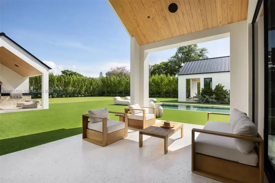 Covered outdoor living area with a natural wood-paneled ceiling, light-toned paved flooring, a rectangular swimming pool, and professional landscaping