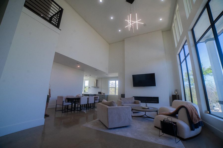 This photo showcases a modern, open-concept living area with high ceilings and large windows, allowing for ample natural light. The space features a sleek fireplace, contemporary furniture, and a minimalist aesthetic, offering a spacious and inviting atmosphere.