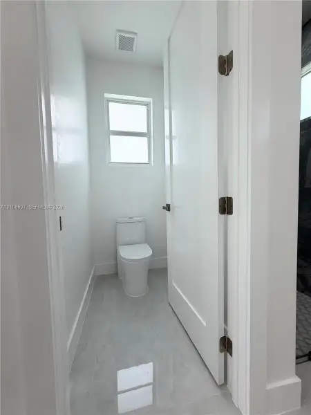 Elongated white toilet, large-format neutral tile flooring, and a single-panel interior door with dark metal hardware