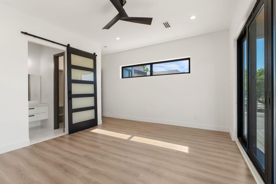 A black barn door with frosted glass panels leads to the en-suite bath on the far wall, while a sliding glass door on the adjacent wall provides a second point of direct access to the rooftop deck. A narrow horizontal clerestory window at the rear adds a layer of natural light without sacrificing privacy.
