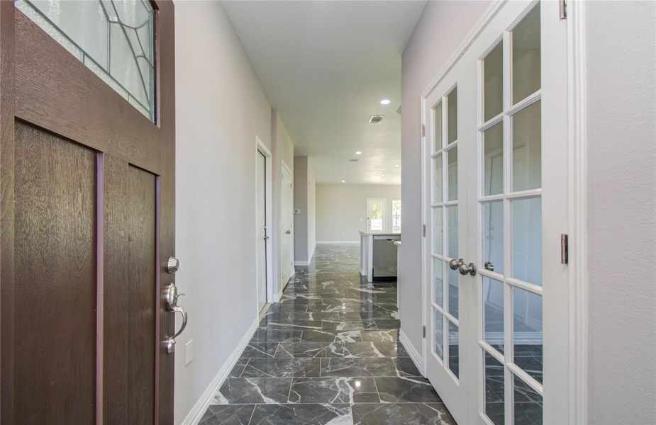 This hallway features elegant dark marble flooring, a wooden front door with decorative glass, and a set of French doors leading to a bright, open living space.