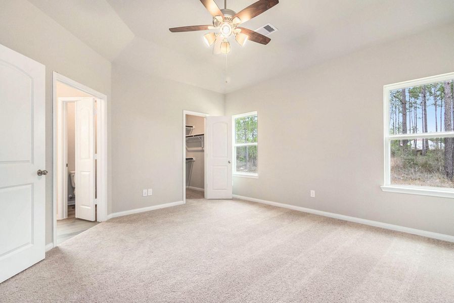 This bright bedroom features neutral walls, carpeted flooring, a ceiling fan, and ample natural light from two windows. It includes an en-suite bathroom and a walk-in closet.