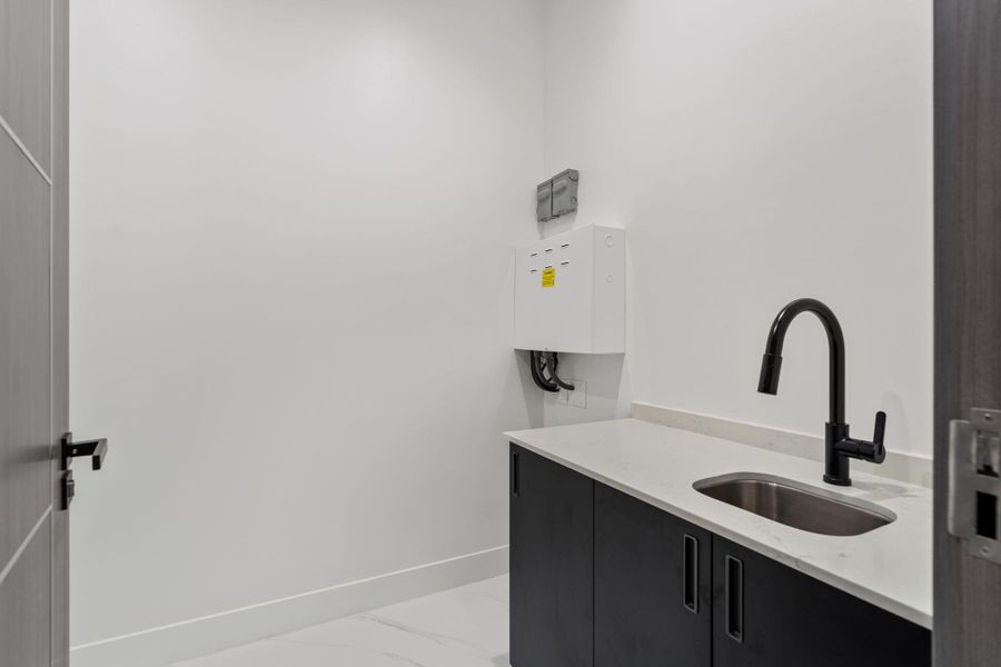 The utility room features dark lower cabinetry below a white countertop with an undermount stainless sink and matte black pull-down faucet, with washer and dryer connections at the far wall. Functional, clean, and thoughtfully equipped.