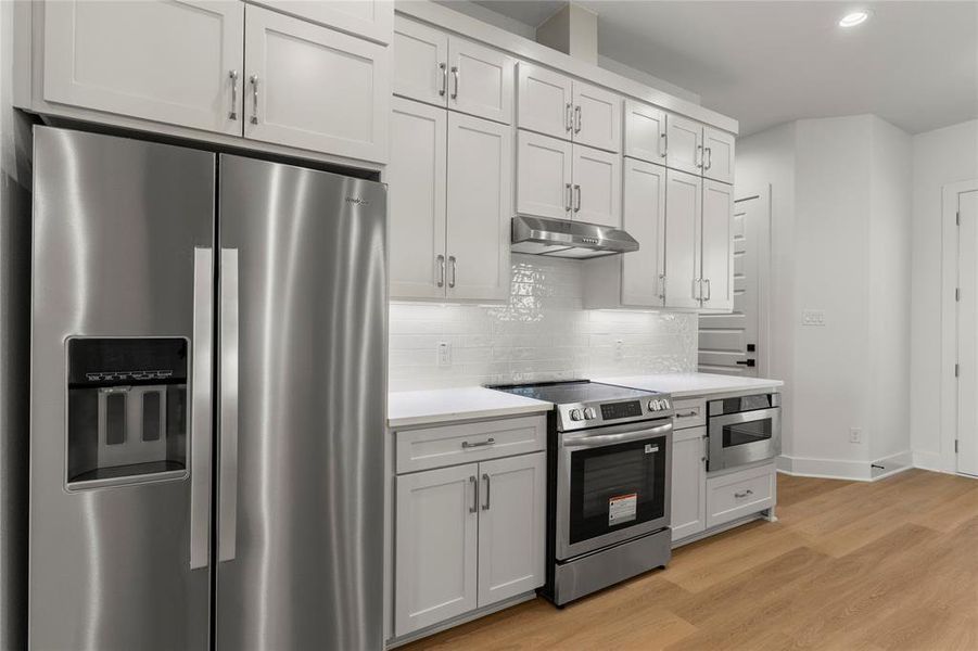 Kitchen with appliances with stainless steel finishes, light wood-style flooring, backsplash, under cabinet range hood, and white cabinetry Kitchen with appliances with stainless steel finishes, light wood-style flooring, backsplash, under cabinet range hood, and white cabinetry