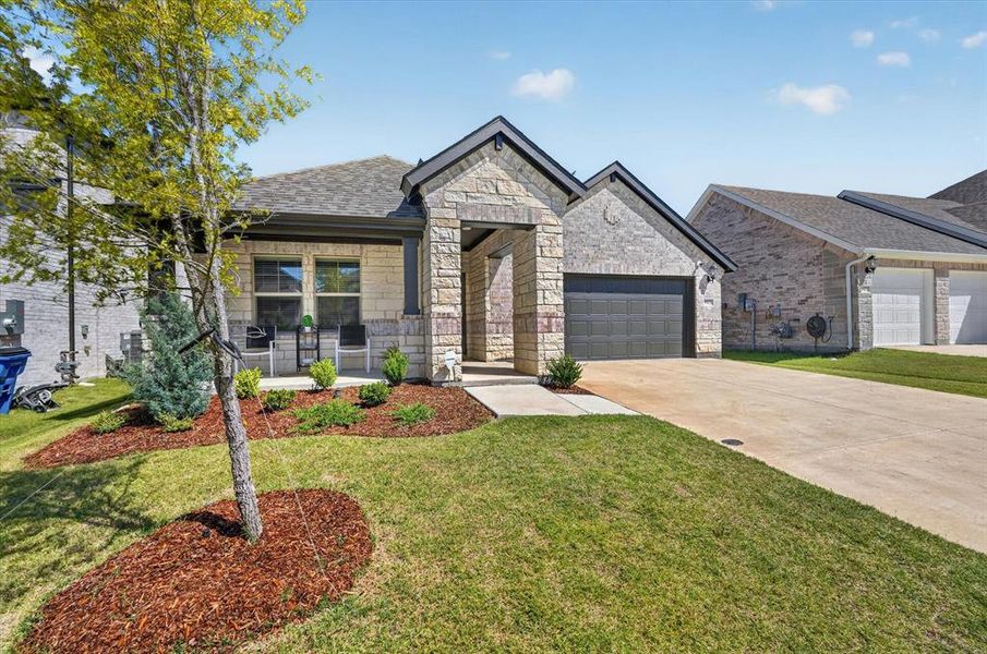 Single-story residence featuring a stone and brick exterior, an attached garage with a dark panel door, a covered porch, a concrete driveway, and landscaped front yard with mulch beds Single-story residence featuring a stone and brick exterior, an attached garage with a dark panel door, a covered porch, a concrete driveway, and landscaped front yard with mulch beds