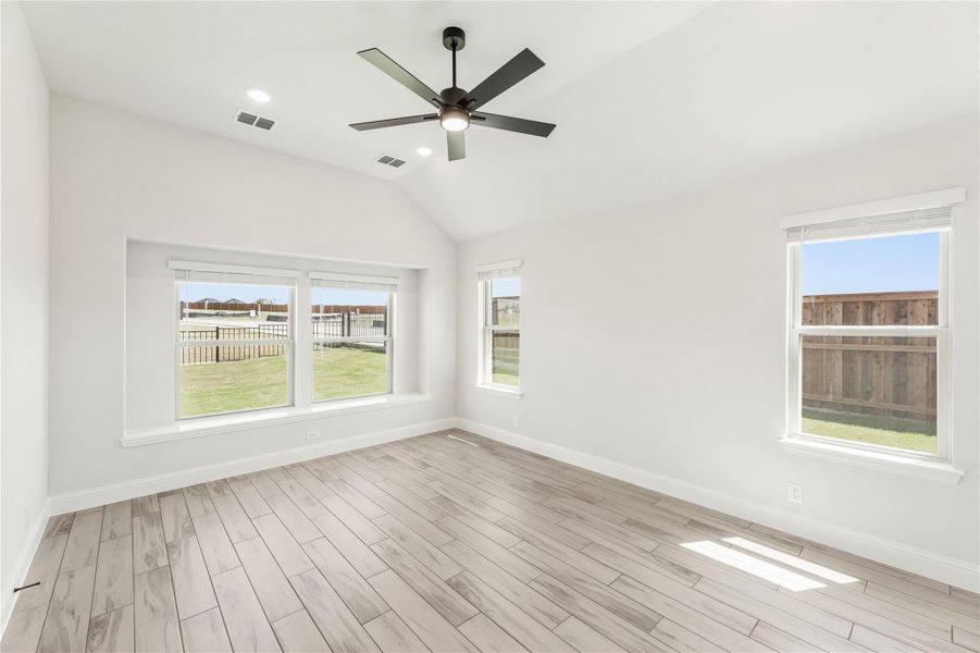 Empty room with vaulted ceiling, ceiling fan, light wood floors, and multiple windows overlooking a backyard