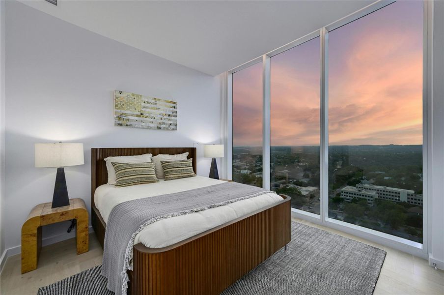 Serene guest bedroom with mesmerizing sunset views. Floor-to-ceiling windows frame a spectacular panorama of the surrounding landscape, creating a captivating backdrop for peaceful evenings. The tranquil space features warm wood accents, soft neutral tones, and thoughtful lighting that complements the natural glow of golden hour, offering the perfect retreat for guests  (Photo from previous model Residence No. 2104 on lower level)