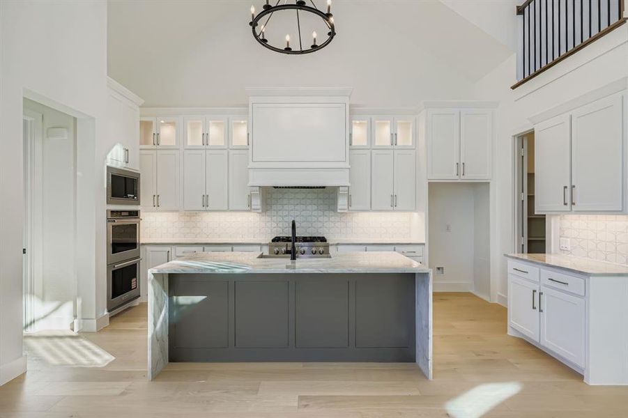 Kitchen with light stone counters, decorative backsplash, white cabinets, high vaulted ceiling, and an island with sink
