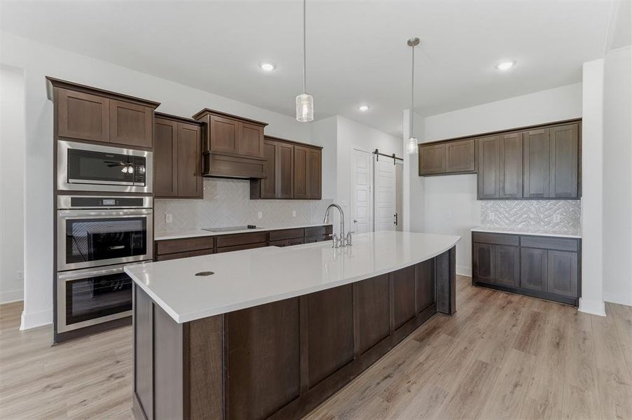Spacious kitchen featuring an expansive island with a white countertop and integrated sink, rich wood-finish cabinetry, stainless steel double ovens and microwave, a patterned tile backsplash, and light wood-finish flooring