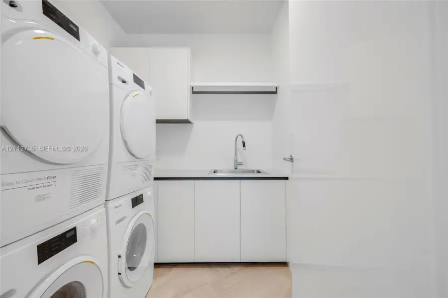 Utility area featuring stacked laundry appliances, white built-in cabinetry, a rectangular stainless steel sink with a high-arch pull-down faucet, and light-tone wood-finish flooring