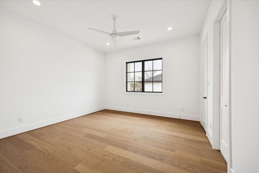 Bright secondary bedroom with wide plank hardwood floors and a black framed window that brings in natural light. Clean white walls, recessed lighting, and a modern ceiling fan create a comfortable, flexible space ideal for family, guests, or a dedicated workspace.