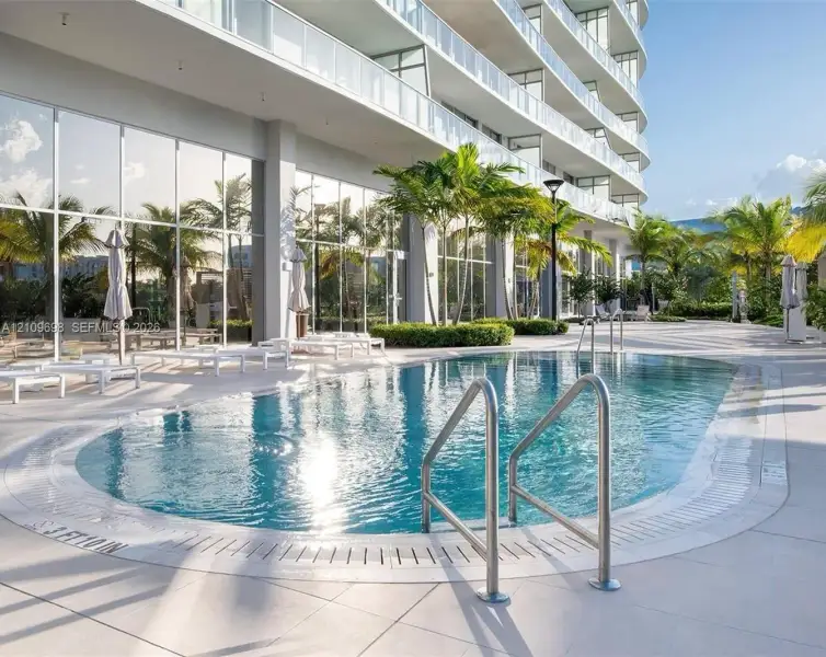 Swimming pool with metal handrails, expansive tile pool deck, floor-to-ceiling glass wall facade, and tropical landscaping