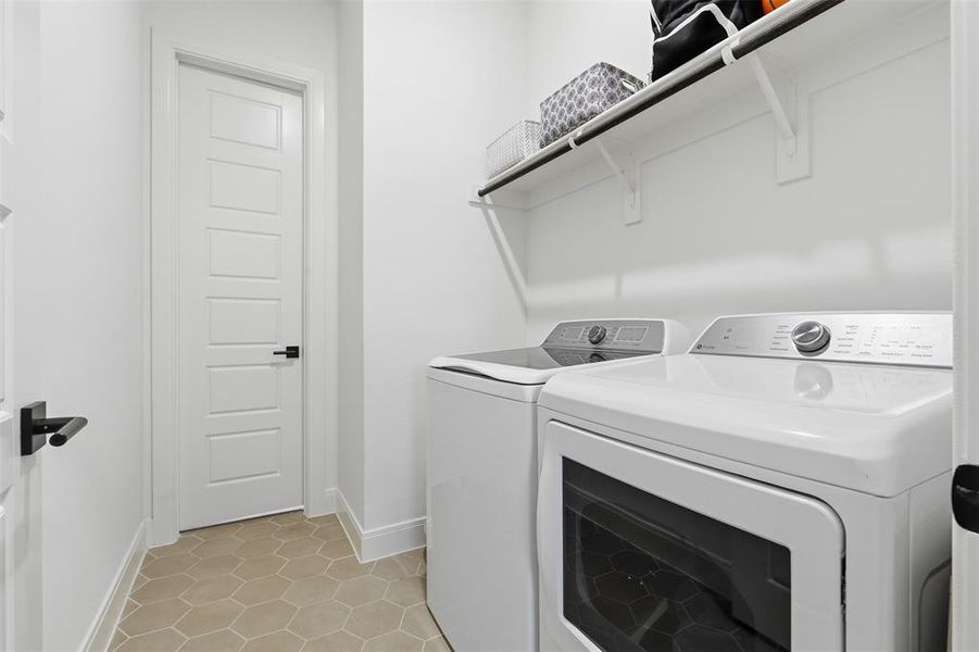 Laundry area featuring hexagon tile flooring, white shaker-style doors with matte black hardware, and an overhead storage shelf Laundry area featuring hexagon tile flooring, white shaker-style doors with matte black hardware, and an overhead storage shelf
