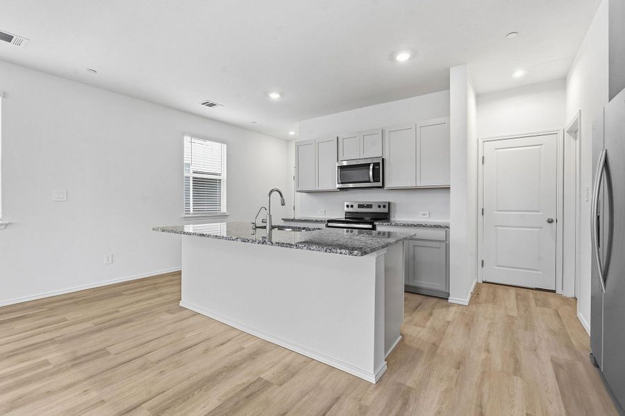 Modern kitchen featuring an island with a sink, stainless steel appliances, light gray cabinetry, speckled countertops, and wood-finish flooring Modern kitchen featuring an island with a sink, stainless steel appliances, light gray cabinetry, speckled countertops, and wood-finish flooring