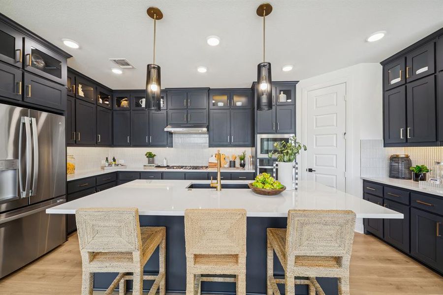 Modern kitchen with dark cabinetry, stainless steel appliances, a large central island with seating, pendant lighting, and light wood-finish flooring